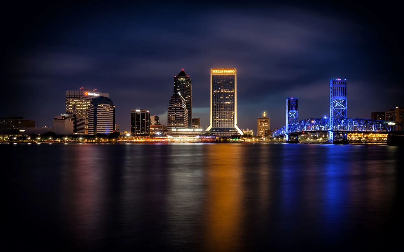 Jacksonville skyline at night across the St. Johns River, with the illuminated Main Street Bridge and downtown towers reflecting on the water.
